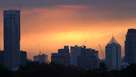 Time-Lapse-Golden-Sunset-Behind-Clouds,-City-Skyscrapers-Skyline-In-Silhouette
