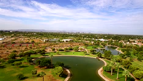 Aerial-slow-push-in-from-the-edge-of-a-pond-at-Granada-Park-toward-the-metro-Phoenix,-​skyline