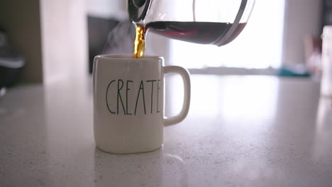 Eine-Kaffeekanne-Gießt-Kaffee-In-Eine-Tasse-Mit-Der-Aufschrift-„Create“,-Bevor-Eine-Hand-Die-Tasse-Aus-Dem-Rahmen-Zieht