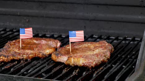 Zwei-Saftige-Rib-Eye-Steaks-Sitzen-Auf-Dem-Grill-Und-Kochen-Mit-Zwei-Winzigen-Zähnen-Der-Amerikanischen-Flagge,-Die-In-Sie-Gestochen-Werden