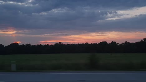 Travelling-and-looking-through-the-window-seeing-sunset,-road-and-forest-with-dark-clouds-and-red-sky