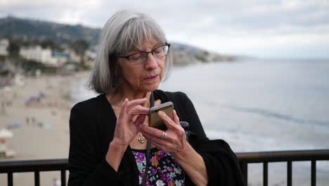 A-middle-aged-woman-with-aging-white-hair-and-eye-glasses-texting-on-her-phone-on-vacation-in-Laguna-Beach,-California-SLOW-MOTION
