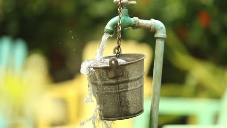 Water-from-a-garden-spout-pours-and-spills-over-a-hanging-bucket-while-the-camera-approaches-with-colored-lounge-chairs-in-the-background