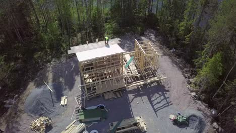 Construction-site-of-a-tiny-country-house-with-beautiful-river-shore,-builder-with-nail-gun-working-on-the-roof,-AERIAL-4K