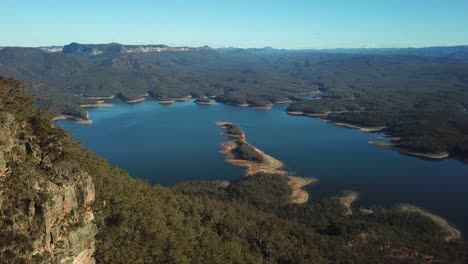 Aerial:-Drone-shot-slowly-tracking-alongside-a-rocky-mountain-towards-a-large-blue-lake-surrounded-by-Australian-bush-land