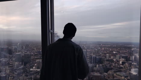 Silhouette-Eines-Erwachsenen-Männlichen-Touristen,-Der-Abends-Aus-Der-Fensterscheibe-Auf-Die-Skyline-Von-London-Blickt