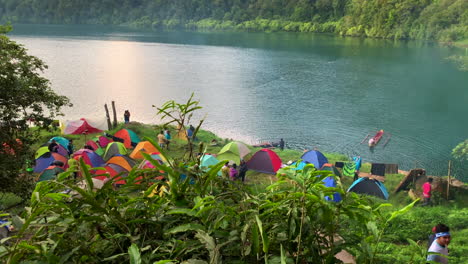 Wide-footage-of-people-camping-by-the-lake-of-Holon,-with-a-colourful-tent-placed-on-the-ground,-a-blue-lake-and-forest-in-the-background