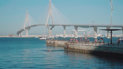 Boy-dives-off-pier-into-bay-with-Cadiz-bridge-in-background,-Spain,-Slowmo