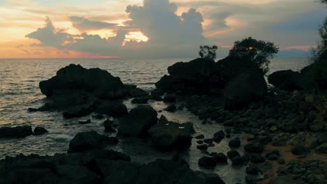 drone-shot-above-ocean-passing-rocky-shore-towards-sunset-behind-the-clouds