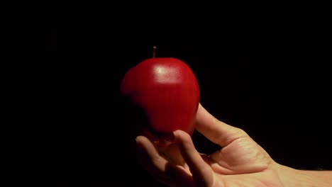 Eine-Weiße-Männliche-Hand-Kommt-Ins-Bild-Und-Hält-Einen-Leuchtend-Roten-Apfel-Vor-Einem-Komplett-Schwarzen-Hintergrund