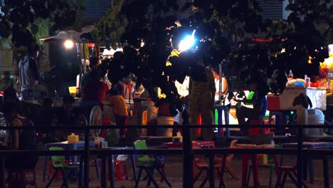 Weitwinkelaufnahme-Eines-Kleinen-Außenrestaurants-Mit-Plastikstühlen-Und-Tischen,-Um-Das-Herum-Menschen-Herumlaufen-Und-Nachts-In-Einer-Hängematte-Schaukeln,-Mit-Verkehr-Im-Hintergrund