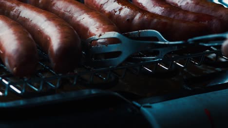 Turning-rotating-fresh-German-sausages-on-electric-BBQ-grill-tongs-green-eco-cooking-global-warming-alternative-Bratwurst-traditional-bavarian-cusine-gloomy-electrical-beef-pork-minced-meat