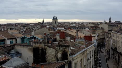 French-city-of-Bordeaux-rooftops-with-Cailhau-City-Gate-and-pigeons-flying,-Aerial-pedestal-lowering-shot