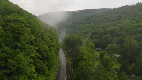 Aerial-drone-flying-overhead-of-curvy-mountain-road-through-green-forest-valley-with-trees-and-rainy-fog
