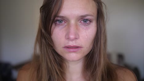 A-close-up,-portrait-shot-of-a-young-woman-with-wounds-and-bruises,-looks-and-stares-directly-into-the-camera-while-a-single-tear-is-falling-down-from-her-eye-onto-her-cheek-and-face