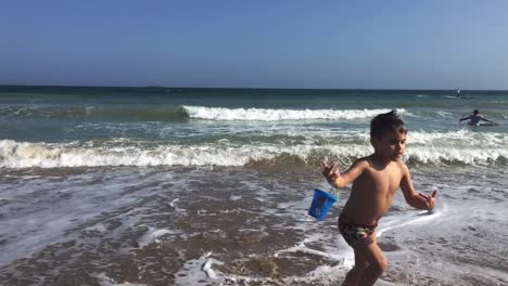 Slow-motion-footage-of-caucasian-toddler-boy,-running-out-of-sea-with-big-waves,-holding-a-toy-bucket-in-his-hands