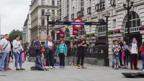 London-underground-sign-above-entrance-to-tube-on-Piccadilly-Circus-square,-people-standing-around-and-watching-street-performance,-outside-wide-angle-view