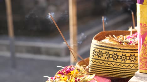 Baskets-filled-with-flowers-for-Hindu-religious-offerings-with-burning-sticks-of-incense-at-temple-in-Bali