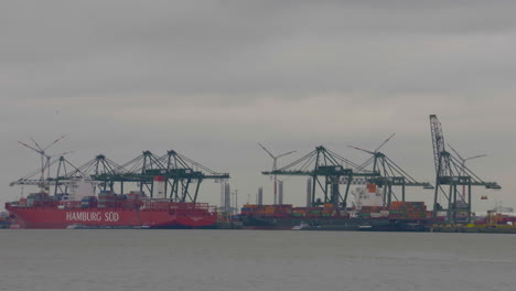 Large-shipping-barges-docked-at-the-Antwerp-Harbor,-loading-and-unloading-shipments-with-windmills-in-the-distance-under-a-cloudy-day---Wide-shot