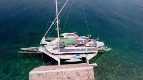 Trimaran-sailboat-Bigkanu-on-an-ocean-reef-dive-cottage,-Aerial-circle-around-shot