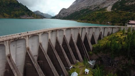 Dam-wall-of-Lake-Fedaia-in-the-Dolomite-mountain-area-of-northern-Italy-with-people-walking-on-structure,-Aerial-Drone-dolly-out-reveal-shot