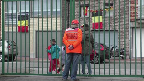 Elderly-man-guarding-gate-as-steward-in-orange-jacket-and-with-orange-cap-at-stadium-before-soccer-game-starts