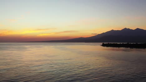 Yellow-clouds-on-bright-sky-after-sunset-reflecting-on-calm-surface-of-seawater-around-silhouette-of-tropical-island-in-Bali