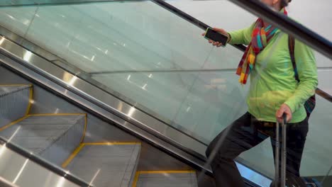Side-shot-closeup-of-woman-going-up-an-escalator-with-smartphone-in-hand-and-roll-on-luggage