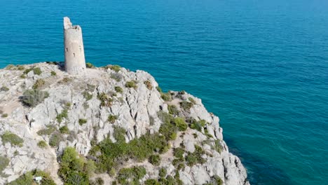 Drone-flies-over-a-coastal-watchtower,-centennial-tower-on-the-blue-sea-cliff-and-rocks-in-a-beautiful-view