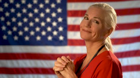 Medium-tight-portrait-of-blonde-nurse-as-she-turns-and-looks-at-camera-looking-relieved-and-happy-with-out-of-focus-US-flag