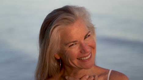 Closeup-of-mature-woman-looking-up-and-then-looking-at-camera-and-smiling-with-out-of-focus-water-behind-her
