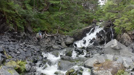 Hiker-Climbing-On-Rocky-Steep-Mountain-With-Cascading-Waterfalls-At-Forest-Park-In-Alaska,-USA