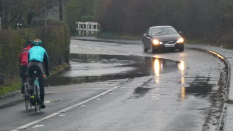 Cyclists-riding-bikes-around-stormy-flash-flooded-road-corner-bend-UK