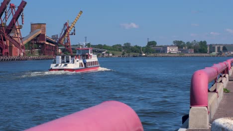 Pleasure-boat-Hercogs-Jēkabs-with-people-on-the-deck-moving-at-Venta-river,-port-cranes-and-harbor-warehouses-in-background,-sunny-summer-day,-Port-of-Ventspils,-wide-shot-from-a-distance