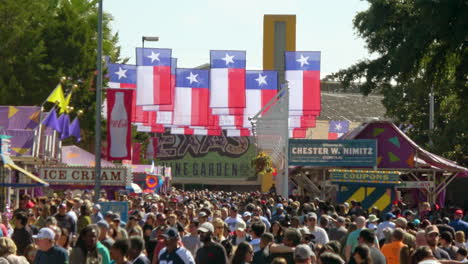 Large-groups-of-people-wander-through-crowded-streets-of-Texas-festival,-Texas-flags-hang-overhead----4K