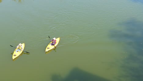 An-aerial-view-of-two-people-slowly-kayaking-from-top-right-to-bottom-left-of-the-screen-on-a-calm,-sunny-day