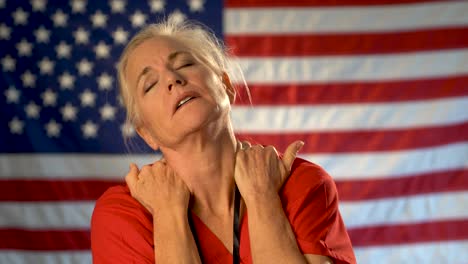 Mittelgroßes-Porträt-Einer-Krankenschwester,-Die-überfordert-Und-Gestresst-Aussieht,-Die-Hände-Im-Nacken-Und-Die-Amerikanische-Flagge-Im-Rücken