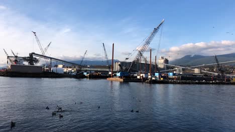 Big-blue-crane-on-a-barge-moored-at-a-pier-with-on-the-background-a-lot-of-grain-conveyors-to-a-silo-while-ducks-are-flying-away-on-the-foreground-on-a-partly-cloudy-day