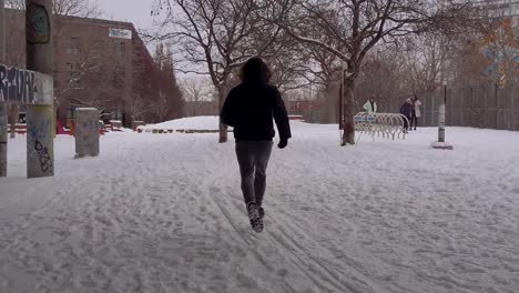 Man-wearing-parka-and-boots-running-on-snow-covered-ground-toward-park-between-buildings-in-Montreal