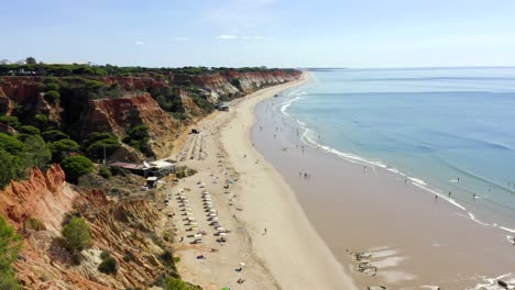 Amazing-golden-beach-and-orange-cliffs-in-Portugal-with-turquoise-waters-and-calm-waves-crashing-in-towards-tourists-enjoying-the-heat