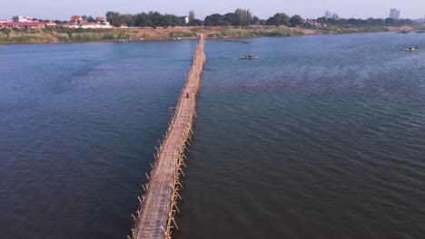 Natural-wooden-bamboo-bridge-with-passing-boat-stretching-over-the-river-Mekong-in-Kampong-Cham,-Cambodia---drone-aerial-fly-over