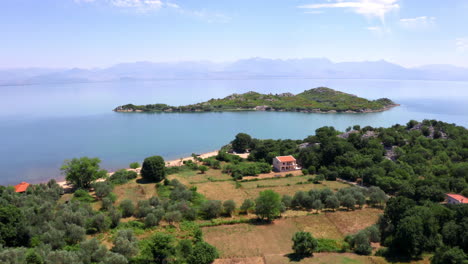 A-small-island-with-lush-vegetation,-crystal-blue-waters-around,-close-to-the-shore-of-lake-Skadar-in-Montenegro-with-houses-and-boats-on-the-beach,-hazy-mountainous-horizon,-aerial-zooming-shot-4K
