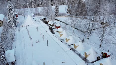 Aerial-view-over-snowy-trees-towards-cottages-and-tents-in-the-Kalevala-Finnish-village,-on-a-overcast-winter-day,-in-Poland---rising,-drone-shot