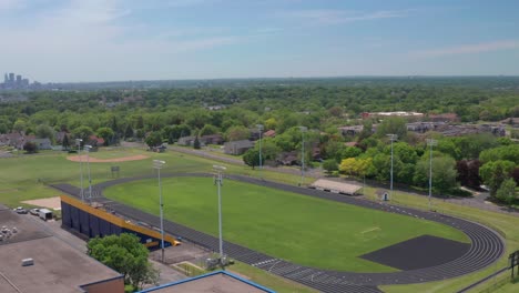 High-school-bleachers-and-track-in-residential-neighborhood-with-city-skyline-on-the-horizon--aerial-drone-flyover-shot