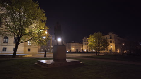 A-statue-of-a-person-on-an-empty-plaza-in-Prague,-Czechia,-at-night,-during-a-Covid-19-lockdown,-trees-and-the-magnificent-concert-hall-of-Rudolfinum-in-the-background,-light-fog-over-the-square,-pan