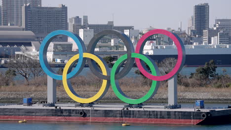Logotipo-De-Los-Juegos-Olímpicos-De-Tokio-2020-Con-Puente-Arcoíris-Y-Paisaje-Urbano-De-Fondo-En-El-Parque-Marino-De-Odaiba-En-Minato,-Tokio,-Japón