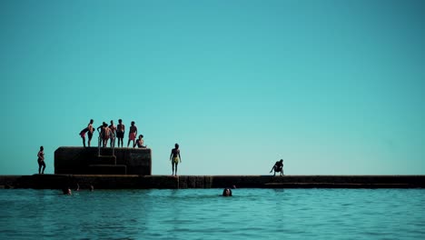 Salzwasser-Swimmingpool-An-Der-Küste-Mit-Kindern,-Die-Bei-Sonnenschein-Vom-Pier-Auf-Das-Wasser-Springen