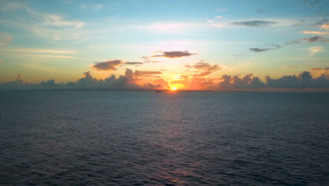 Spectacular-timelapse-view-of-the-sun-rising-from-beyond-the-Caribbean-horizon,-blue-sky-above,-clouds-resembling-a-city-skyline,-a-distant-cruise-ship-passing-the-sun-disc,-scenic-timelapse-FHD-shot