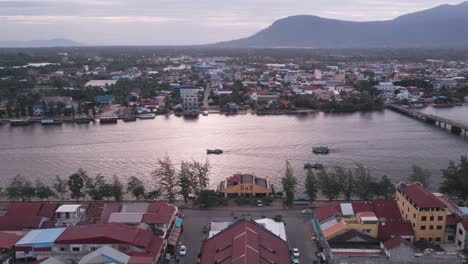 Kampot-town,-old-market-drone-lift-shot-looking-towards-Bokor-hill-and-fishing-boats-traveling-along-the-river