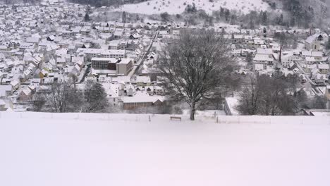 Dronie-shot,-flaying-backwards-from-a-snow-covered-tree-with-a-bench-back-to-dicover-a-valley-with-a-snow-covered,-white-and-dreamy-town